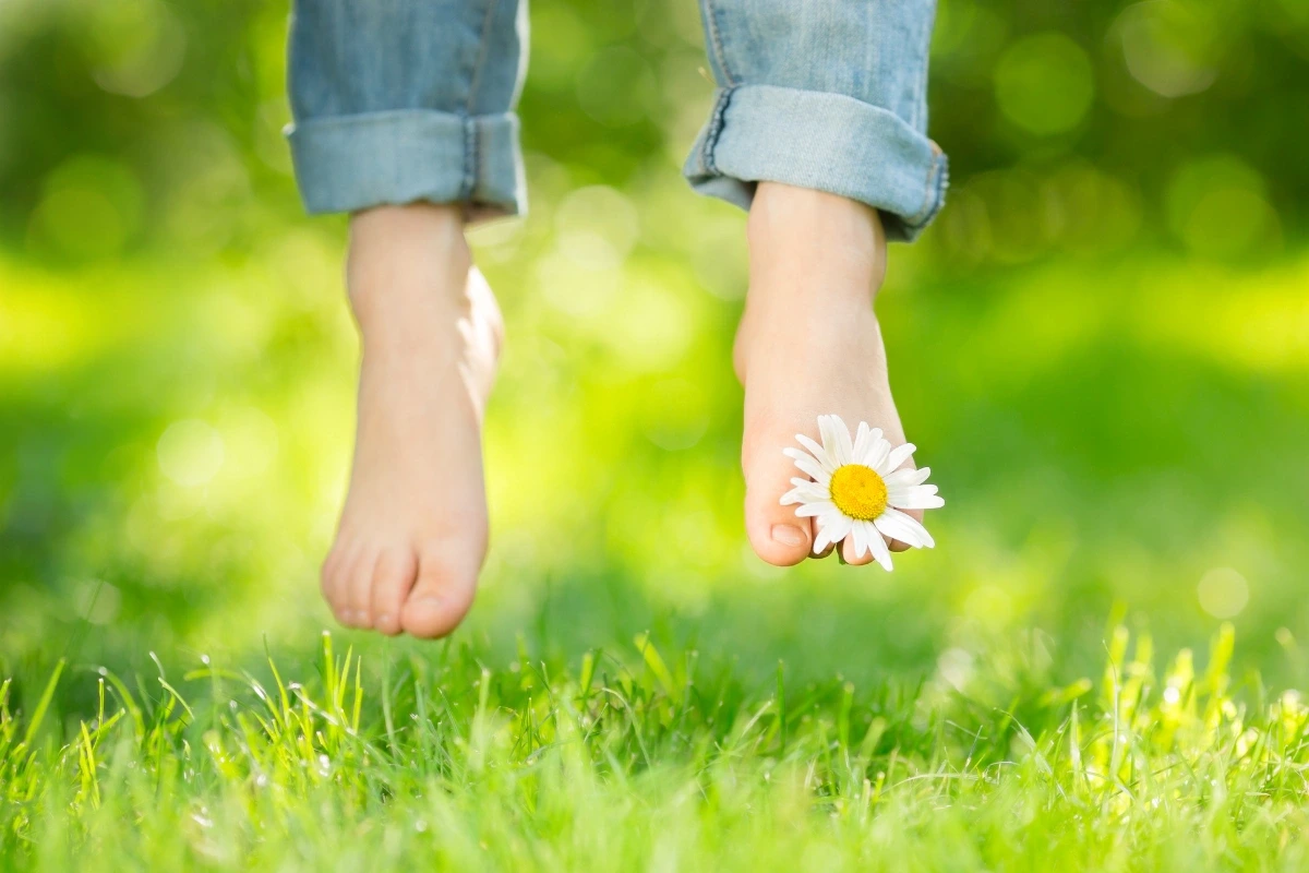 Bosé nohy so sedmokráskou na palci visiace nad svetlozelenou trávou.