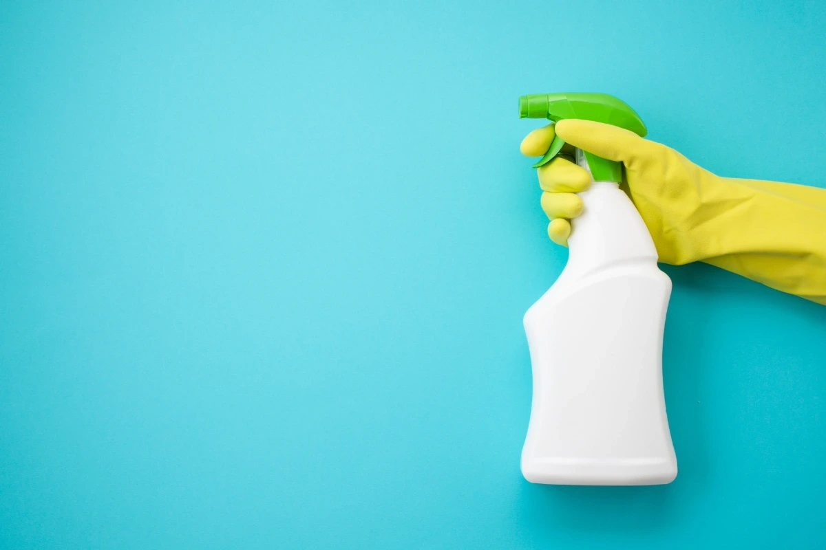 Ruka v žltej gumenej rukavici drží biely rozprašovač so zelenou hlavicou na modrom pozadí.