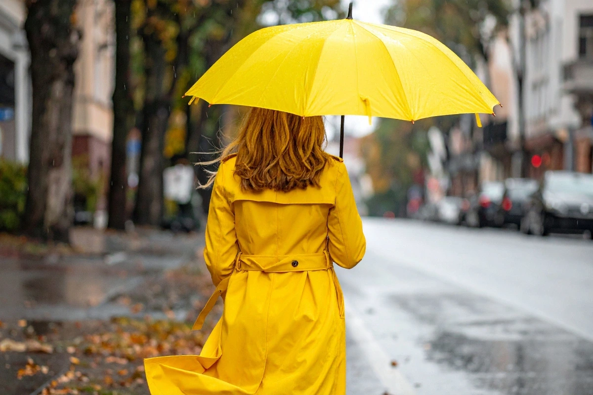 Žena v žltom pršiplášti a so žltým dáždnikom kráča chrbtom k fotoaparátu po mokrej ulici s jesennými listami.