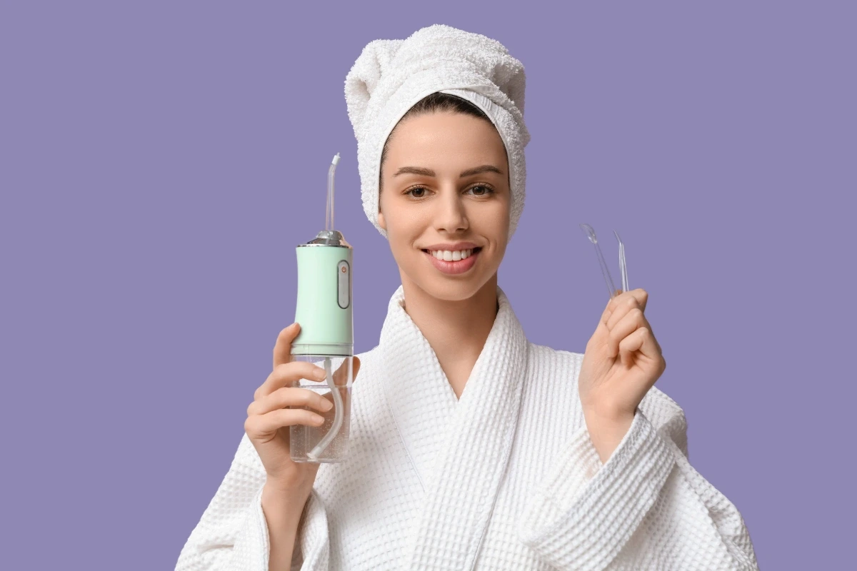 Žena v bielom župane a s uterákom na hlave drží ústnu sprchu a náhradné trysky.