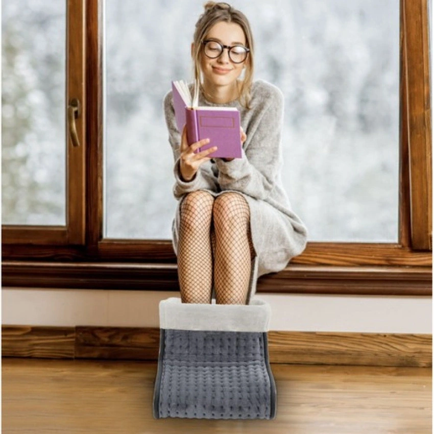 Mladá žena sedí s knihou pri okne s nohami v elektrickom vyhrievači na nohy.