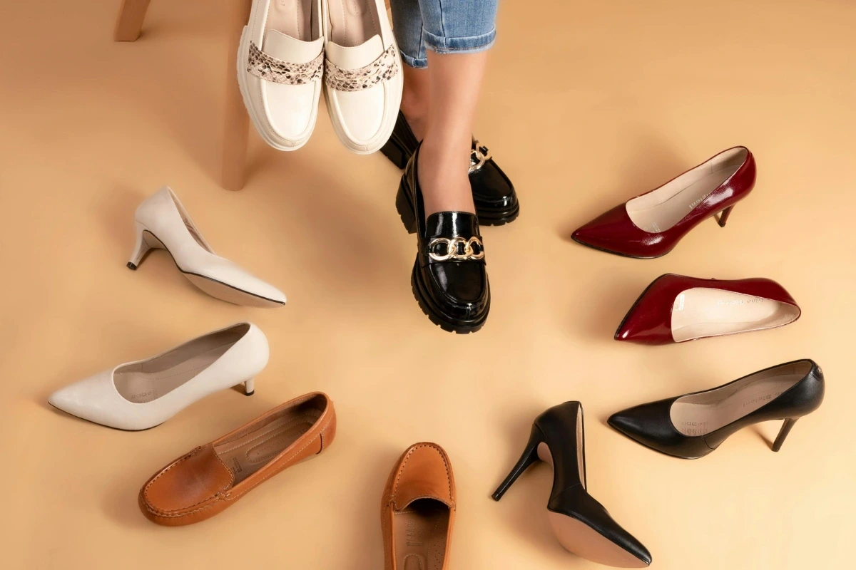 Záber na ženské nohy, okolo ktorých sú rozmiestnené rôzne typy dámskej obuvi: lodičky s úzkou špičkou aj širšie poltopánky. 