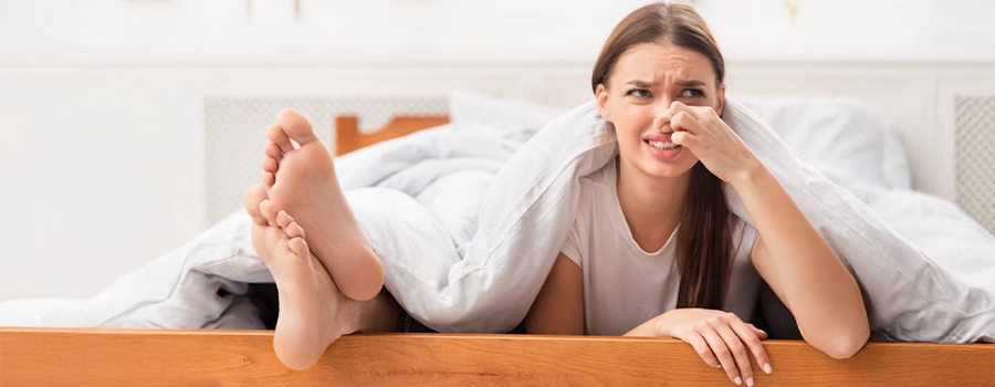 Žena leží vedľa nôh vytŕčajúcich spod periny a znechutene si zakrýva nos kvôli zápachu.